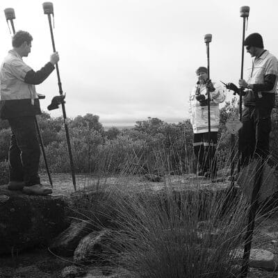 black & white image of three male surveyors holding range poles with GPS. They are standing on a rock in bushland