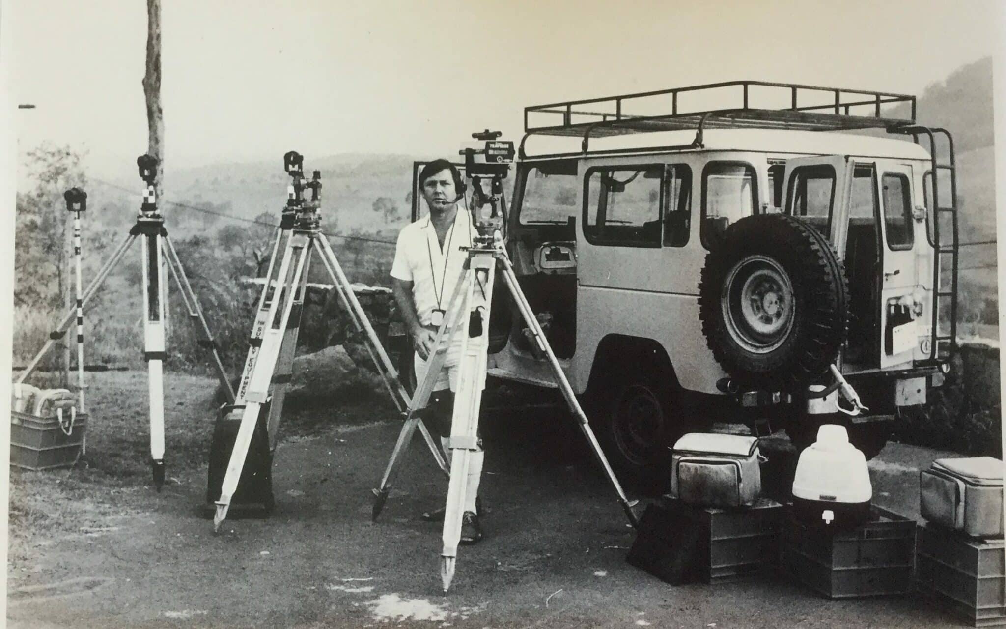 male standing behind several tripods with various survey equipment attached, taken outdoors beside a 4WD. Black & White image